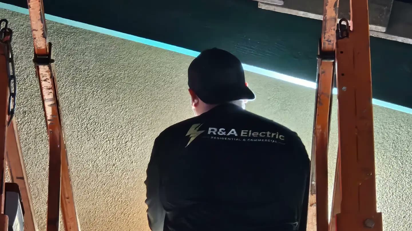 An R&A Electric electrician on a scissor lift at night, his back to the camera with the company logo across his shirt, working under a newly replaced wall pack light.