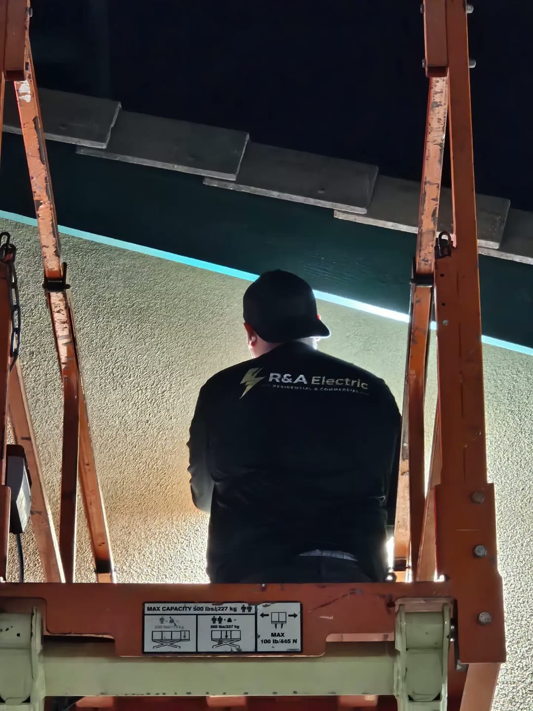 The back of an R&A Electric crew shirt, the gold bolt logo across the shoulders, on a scissor lift under a wall pack.