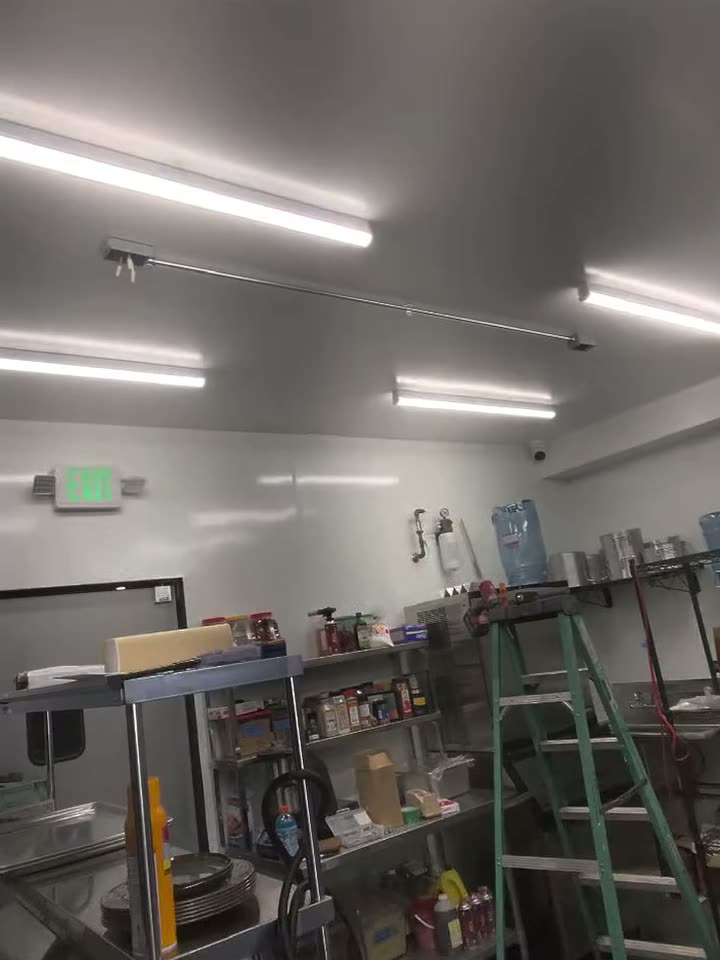A commercial kitchen lit by long LED strip lights, an exit sign on the far wall and stainless shelving below.