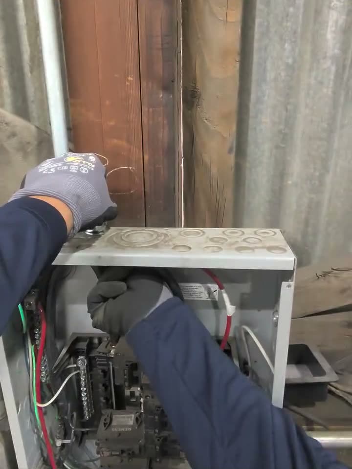 Gloved hands terminating wires in a box at the end of an EMT conduit run on a corrugated shop wall.