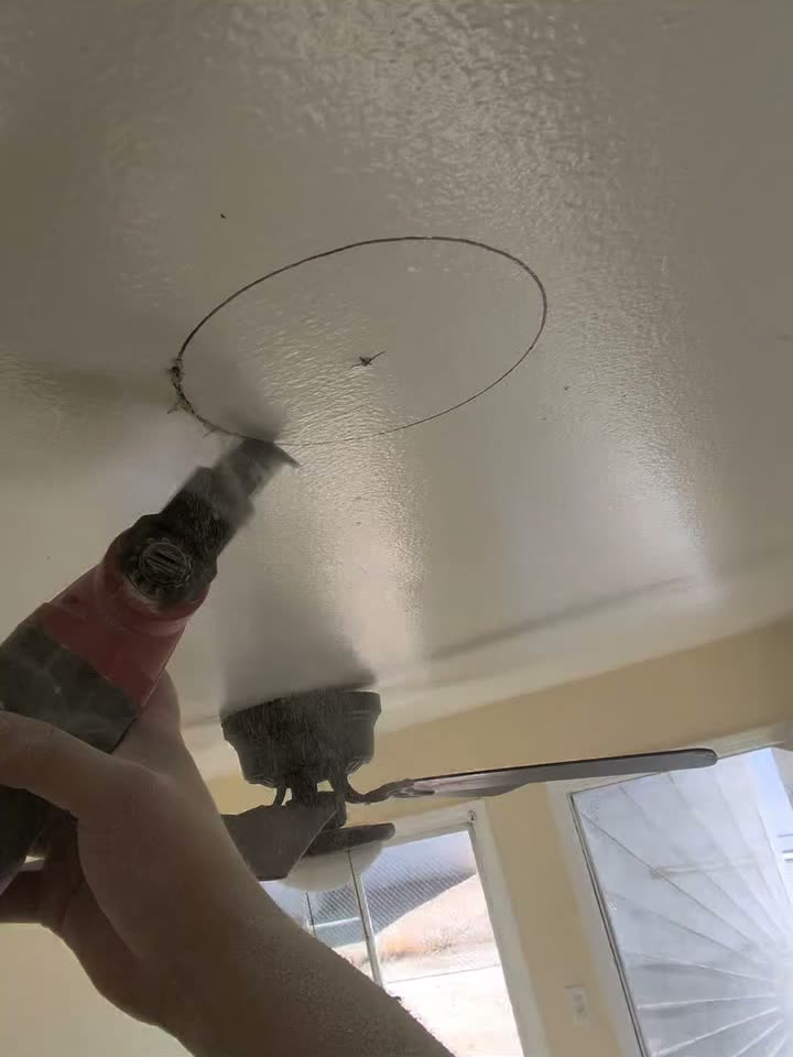 An electrician cutting a round hole in a textured ceiling with a drill, the circle for a recessed light marked out above him.